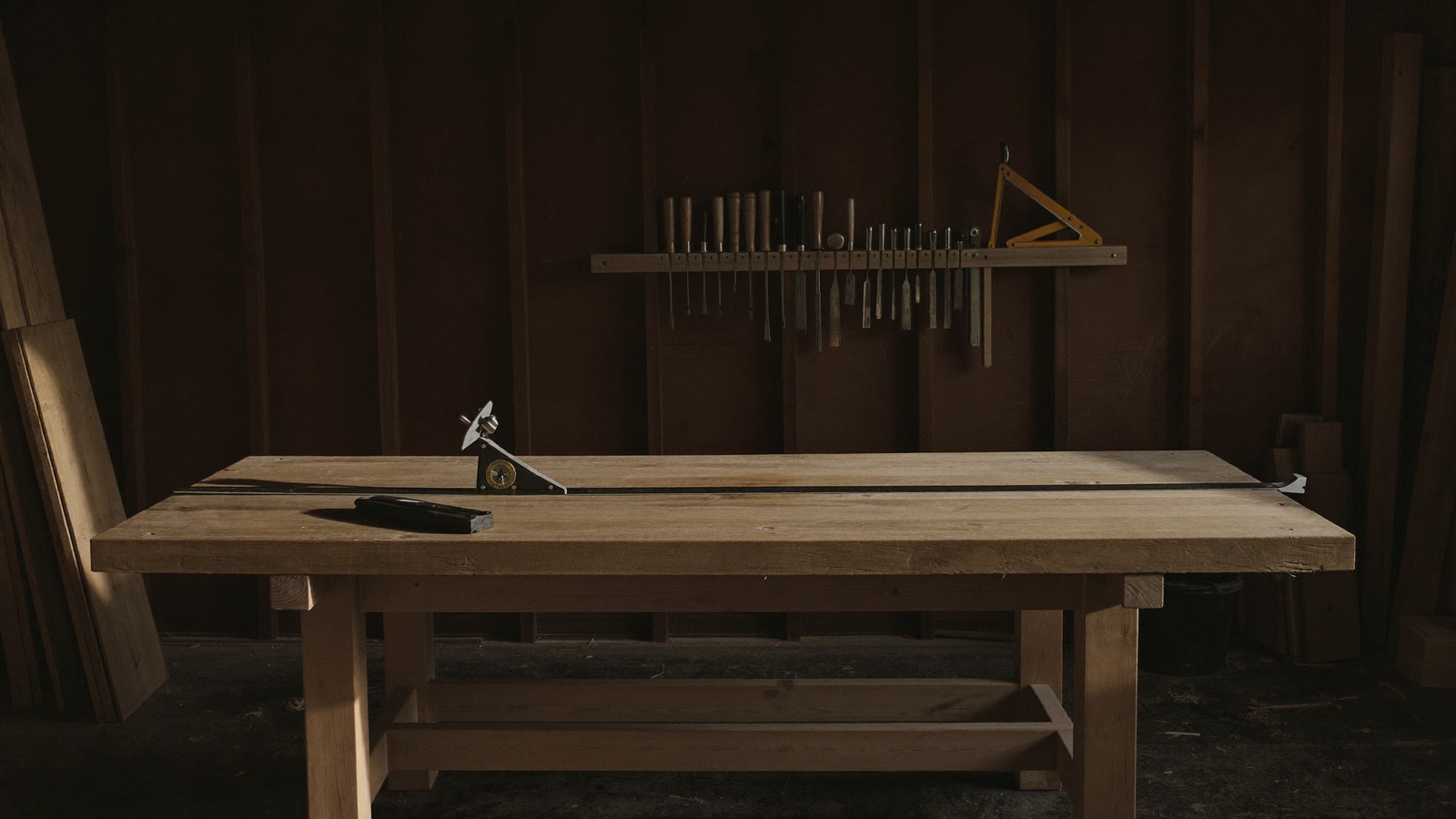 An empty workbench in the workshop, swept clean, a straight edge and a marking gauge lying on it and hand tools racked on the dark wall behind.